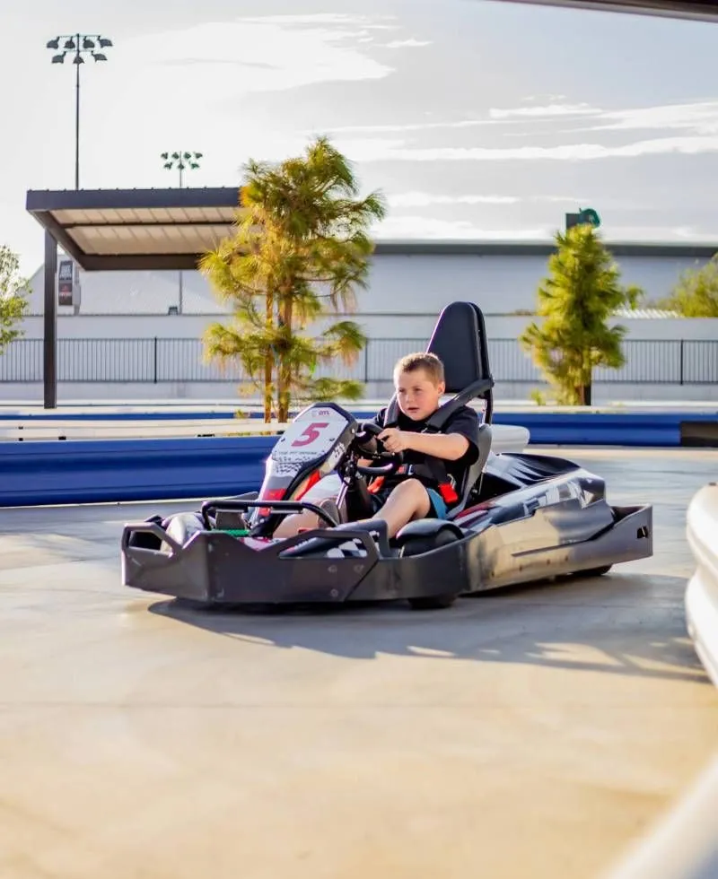 Child driving a black go-kart on an outdoor track in bright daylight