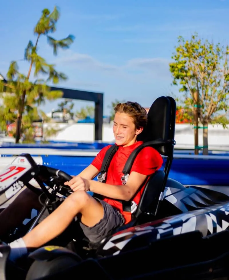 Person seated in a red kart, smiling on an outdoor track with blue sky and trees in the background