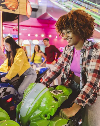 People riding colorful arcade motorcycles indoors, with a smiling rider in front on a green bike.