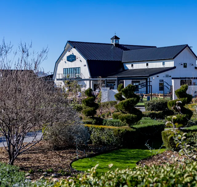 White farmhouse with dark roof behind manicured garden and trimmed shrubs under a clear blue sky